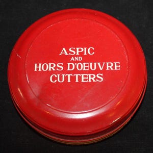 May include: A round, red container with the words "ASPIC AND HORS D'OEUVRE CUTTERS" in white lettering. The container appears to be made of metal and has a slightly worn appearance. The lid is slightly domed.