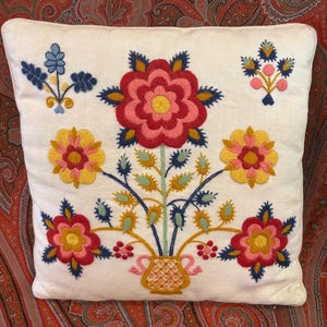 May include: A square white throw pillow featuring a central embroidered design of a yellow vase holding a large red flower, flanked by two yellow flowers and two smaller red flowers, with blue and green accents and small floral sprigs.