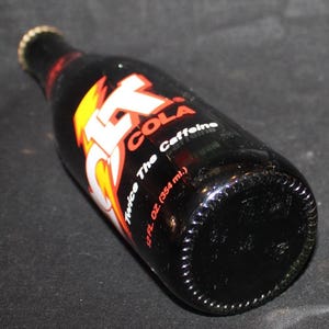 May include: A black glass bottle of Jolt Cola, with a red, white, and yellow logo. The bottle has a gold cap and the label reads "Twice The Caffeine" and "12 fl. oz. (354 ml.)".