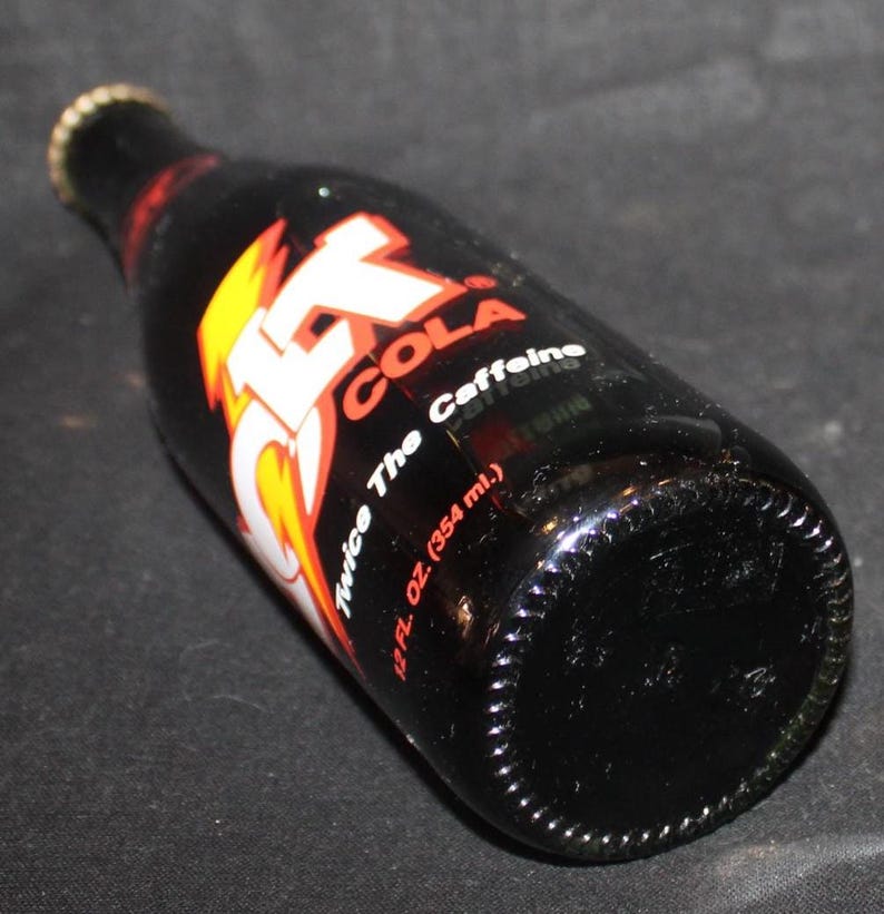May include: A black glass bottle of Jolt Cola, with a red, white, and yellow logo. The bottle has a gold cap and the label reads "Twice The Caffeine" and "12 fl. oz. (354 ml.)".