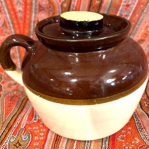 May include: A ceramic crock with a glossy dark brown upper section and a cream-colored lower base. The pot features a matching dark brown lid with a flat, light-colored circular knob on top and a single curved handle extending from the side.