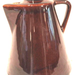 May include: A dark brown ceramic pitcher with a glossy finish, featuring a wide base that tapers toward a narrow top, a single side handle, and a prominent spout. The surface reflects light, highlighting the smooth, glazed texture of the vessel.