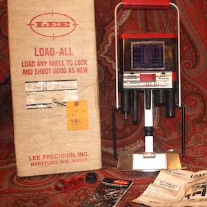 May include: A vintage Lee Load-All reloading tool with its original packaging. The box is beige with red text and a logo. The reloading tool is black, silver, and red. The text on the box reads "LOAD-ALL" and "LOAD ANY SHELL TO LOOK AND SHOOT GOOD AS NEW."