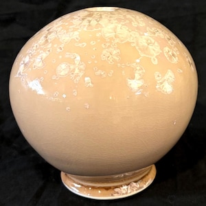 May include: A spherical ceramic vase with a glossy peach-colored glaze featuring white crystalline patterns across the surface. The vase has a small circular opening at the top and a flared base, with a visible fine crackle texture throughout the finish.