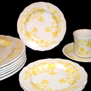 May include: White ceramic dinnerware set with a yellow floral design. The set includes plates, saucers, and a teacup. The plates are stacked, and the teacup is on a saucer. The floral pattern is repeated on each piece.