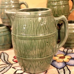 May include: A collection of green ceramic pitchers and mugs. The pitcher has a spout and handle, and the mugs have handles. The pieces have a barrel-like design with vertical lines and horizontal bands. The items are on a floral patterned tablecloth.