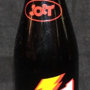 May include: A vintage Jolt Cola bottle with a gold cap. The black bottle features the Jolt Cola logo in red and white with a yellow lightning bolt. The label states "Twice The Caffeine" and the volume is 12 fl. oz. (354 ml).