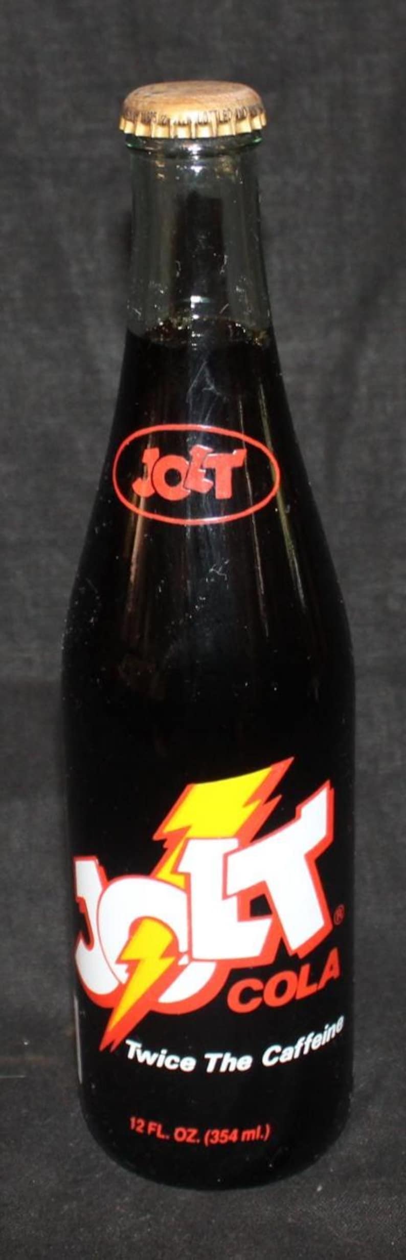 May include: A vintage Jolt Cola bottle with a gold cap. The black bottle features the Jolt Cola logo in red and white with a yellow lightning bolt. The label states "Twice The Caffeine" and the volume is 12 fl. oz. (354 ml).