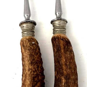 May include: Two silver-toned metal forks featuring four tines each. The handles are crafted from textured brown antler material, accented with decorative silver-toned metal ferrules at the top and bottom of each handle, providing a distinct look.
