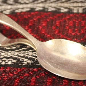May include: A silver baby spoon with a curved handle. The spoon is lying on a red and black patterned fabric.