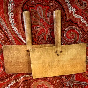 May include: Two rectangular wooden paddles with vertical handles attached to the center of each flat blade. The light brown wood shows a natural grain and texture, with three small metal nails visible at the base of each handle connection.