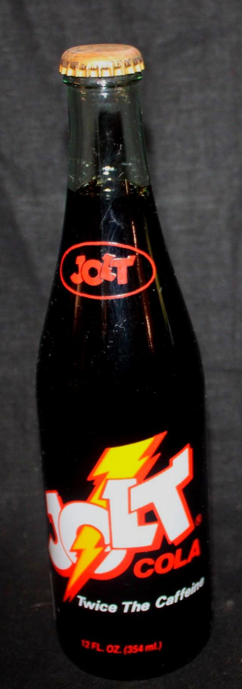 May include: A vintage Jolt Cola bottle with a black body and a gold cap. The label features the Jolt logo in red and white with a yellow lightning bolt. The bottle contains 12 fl oz (354 ml) of cola.