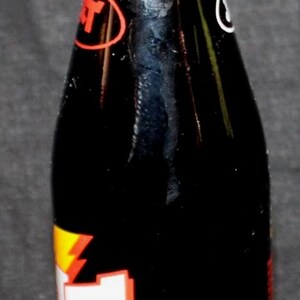 May include: A vintage glass bottle of Jolt Cola, a caffeinated soft drink. The bottle is mostly black with red, white, and yellow branding. The bottle cap is gold-colored.