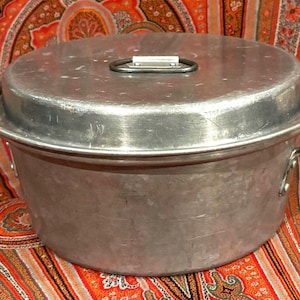 May include: A round, brushed aluminum pot with a matching lid. The lid features a central metal handle with a white plastic grip. The pot has two side handles made of dark metal, securely attached with rivets to the main body of the cookware.