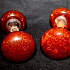 May include: Four round, reddish-brown bottle stoppers with a glossy finish. The stoppers have a marbled appearance and are attached to clear glass stems. The background is black.