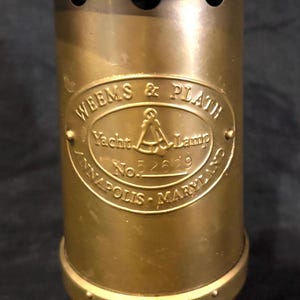 May include: A vintage brass yacht lamp with the text "WEEMS & PLATH Yacht Lamp Annapolis Maryland No. 52629" on the side. The lamp has a cylindrical body, a domed top with air vents, and a carrying handle. The lamp is made of brass.