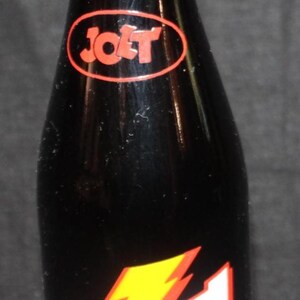 May include: A vintage Jolt Cola bottle with a black background. The bottle has a red and yellow lightning bolt logo and the text "Jolt Cola Twice The Caffeine". It contains 12 fl oz (354 ml).