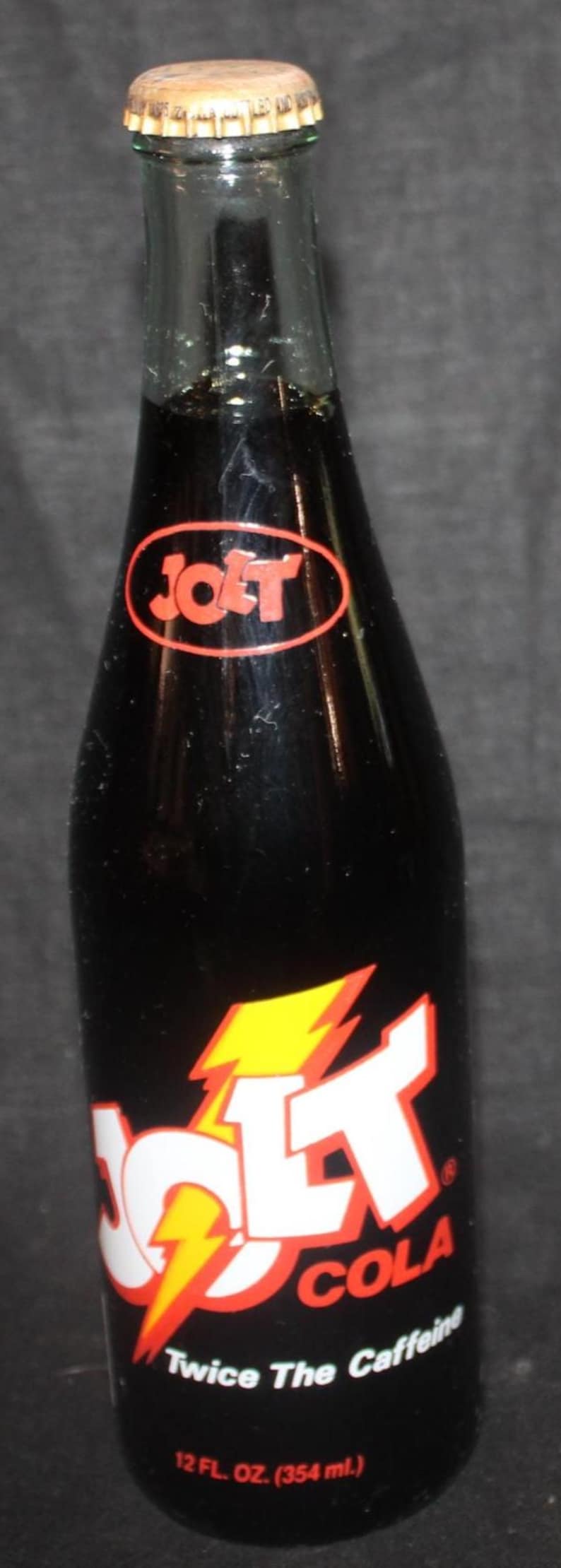 May include: A vintage Jolt Cola bottle with a black background. The bottle has a red and yellow lightning bolt logo and the text "Jolt Cola Twice The Caffeine". It contains 12 fl oz (354 ml).
