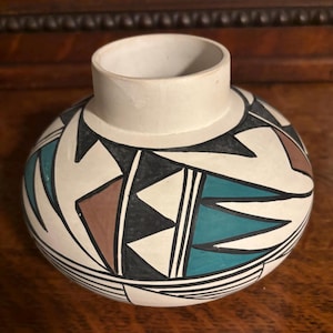 May include: A round, off-white ceramic vase with a short neck. The vase is decorated with geometric patterns in black, teal, brown, and white. The design includes triangles and lines. The vase sits on a wooden surface.