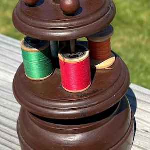 May include: A vintage wooden thread holder with multiple levels. The top level has several small, round knobs. The middle level holds spools of green, red, and orange thread. The thread holder is brown and sits on a wooden surface.