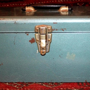 May include: A rectangular, teal-colored metal toolbox featuring a black handle on top and a central metal latch. The exterior shows signs of wear with visible paint chips and rust. A small, rectangular logo with red and black text is on the bottom right.