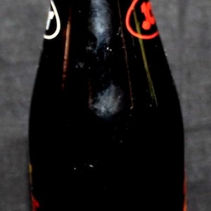 May include: A vintage glass soda bottle with a brown metal cap. The bottle is filled with a dark liquid and has white and red labels with the number "50" prominently displayed. The bottle is against a dark background.