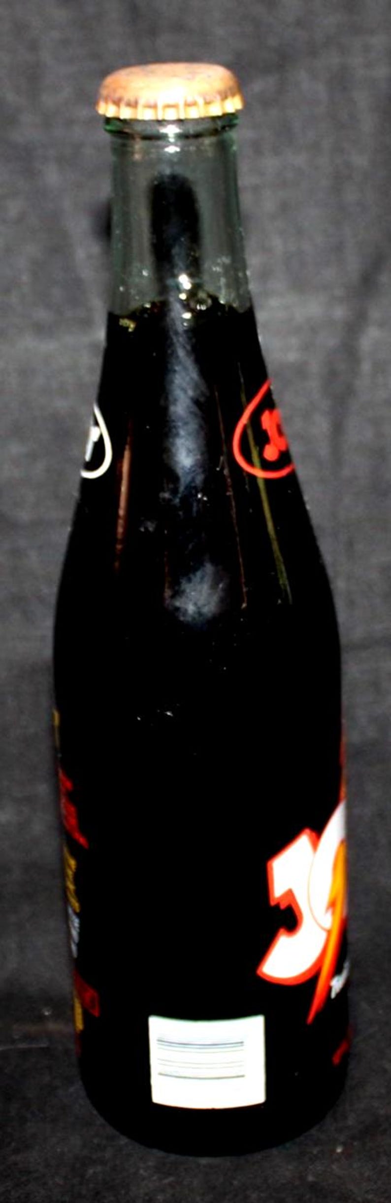 May include: A vintage glass soda bottle with a brown metal cap. The bottle is filled with a dark liquid and has white and red labels with the number "50" prominently displayed. The bottle is against a dark background.
