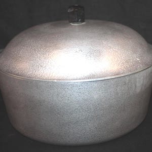 May include: A vintage, silver-colored cooking pot with a lid. The pot has a textured surface and two side handles. The lid is domed with a black knob. This cookware is suitable for various cooking needs.