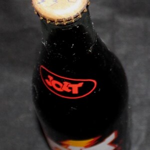 May include: A dark glass bottle of Jolt Cola, with a gold-colored bottle cap. The bottle has a red and white label with the word "Jolt" in a red oval. The bottle cap has the word "Jolt" on it.