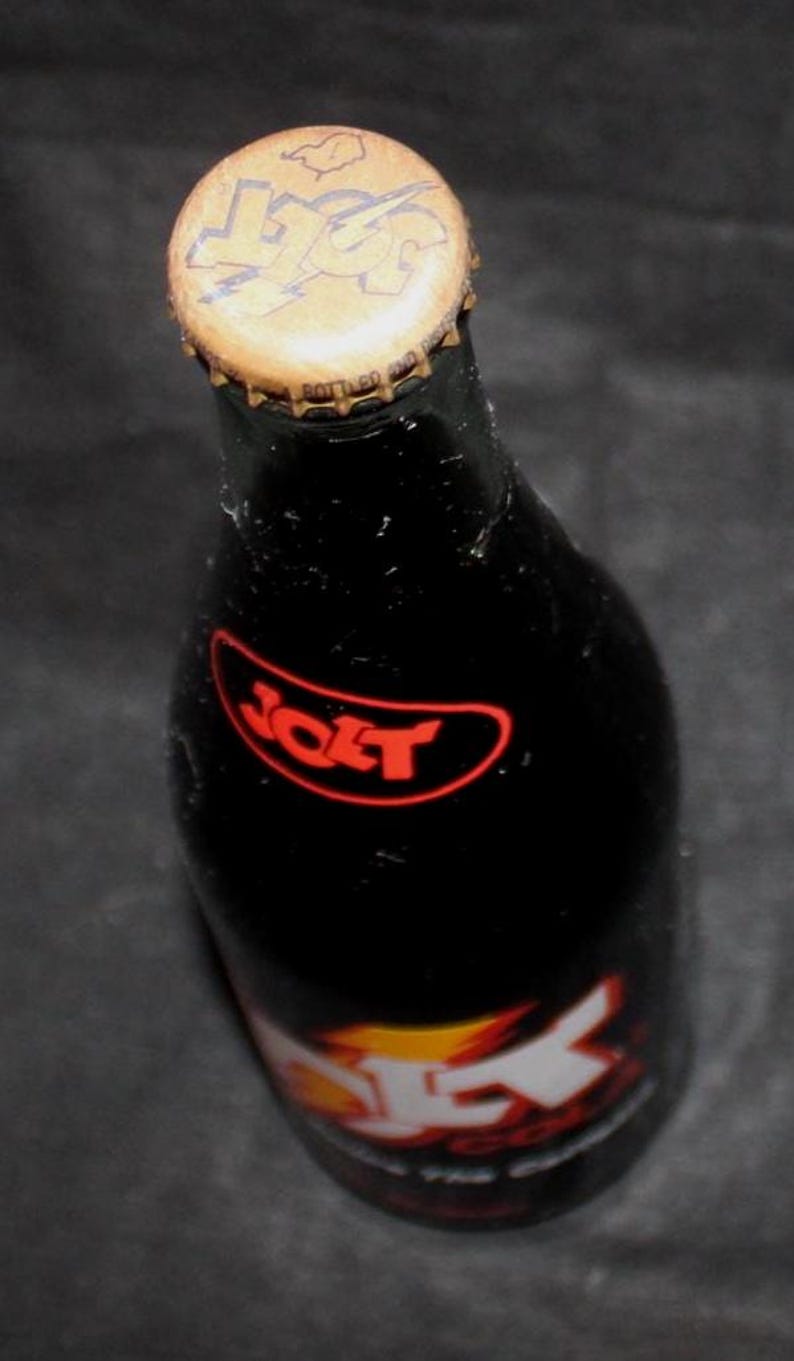 May include: A dark glass bottle of Jolt Cola, with a gold-colored bottle cap. The bottle has a red and white label with the word "Jolt" in a red oval. The bottle cap has the word "Jolt" on it.