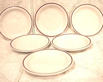 May include: A set of six white ceramic plates with a maroon rim. The plates are arranged on a white lace tablecloth. The set includes both round and oval-shaped plates, suitable for serving food.