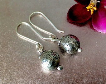 May include: A pair of silver dangle earrings. Each earring features a textured, silver-colored bead suspended from a curved ear wire. The earrings are displayed on a pink surface, with a dark pink flower in the background.