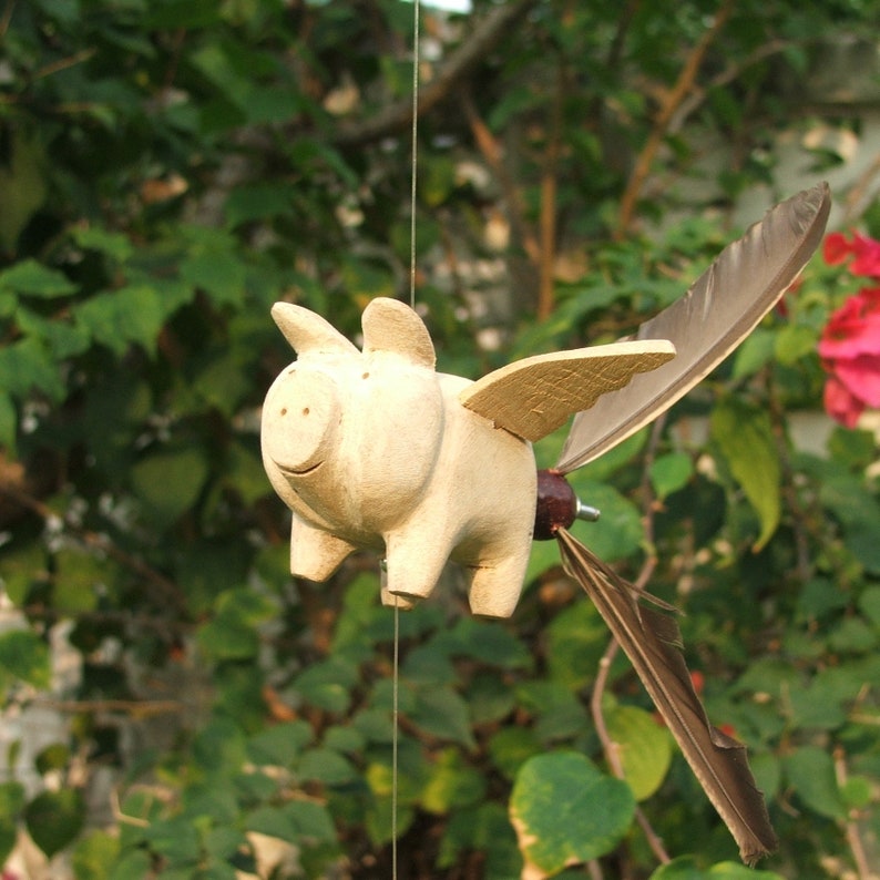 May include: A wooden pig figurine with wings made of feathers. The pig is painted white and has a smiling face. The wings are attached to the pig's back with a small metal screw.