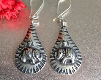 May include: A pair of silver-toned drop earrings. Each earring features a teardrop shape with an embossed elephant design in the center, surrounded by geometric patterns. The earrings have a hook closure. The background is a metallic silver color.