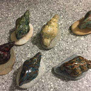 May include: Six ceramic seashells, each with a unique glaze and color, are displayed on small, gray rocks. The shells are in various shades of green, brown, and beige.