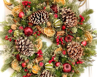 May include: A Christmas wreath with a variety of natural elements, including pine cones, evergreen branches, red berries, dried oranges, and cinnamon sticks. The wreath is decorated with a rustic twine bow and is hanging on a white door.