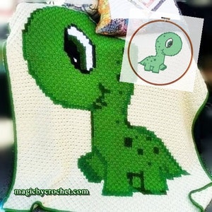 May include: A handmade crochet blanket with a green dinosaur design on a cream background. The dinosaur has a large eye and a long neck. The blanket has a green border. The website magicbycrochet.com is visible. The image also shows a small cross-stitch dinosaur.
