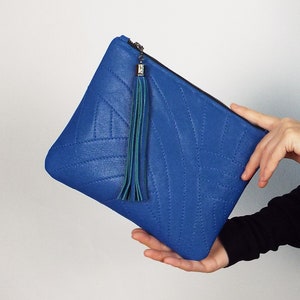 May include: A bright blue clutch bag with a quilted design and a teal tassel. The bag has a zip closure. The clutch is held by a person. The bag is made of a textured material.