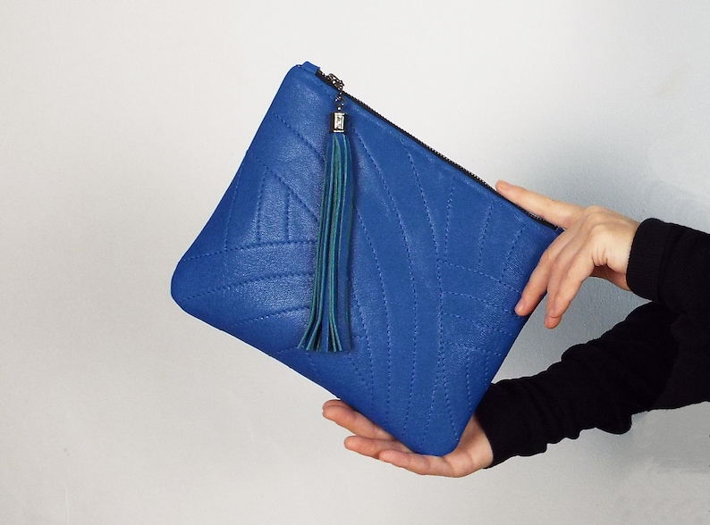 May include: A bright blue clutch bag with a quilted design and a teal tassel. The bag has a zip closure. The clutch is held by a person. The bag is made of a textured material.