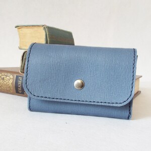 May include: A small, light blue leather wallet with a snap closure and a silver button. The wallet is sitting on top of a stack of books.