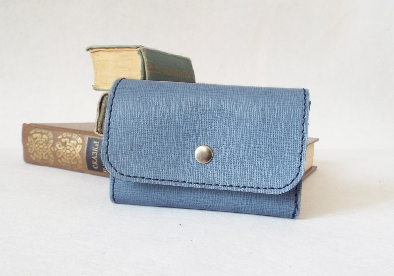 May include: A small, light blue leather wallet with a snap closure and a silver button. The wallet is sitting on top of a stack of books.