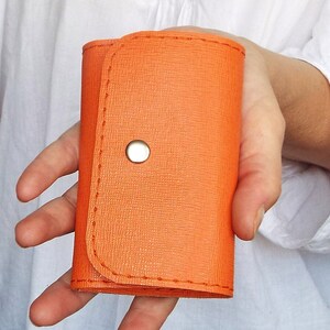 K&ouml;nnte beinhalten: Ein kleiner orangefarbener Schl&uuml;sselanh&auml;nger aus Leder mit einem silbernen Druckknopfverschluss.