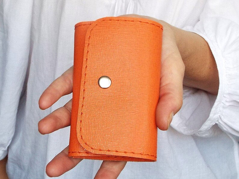 K&ouml;nnte beinhalten: Ein kleiner orangefarbener Schl&uuml;sselanh&auml;nger aus Leder mit einem silbernen Druckknopfverschluss.