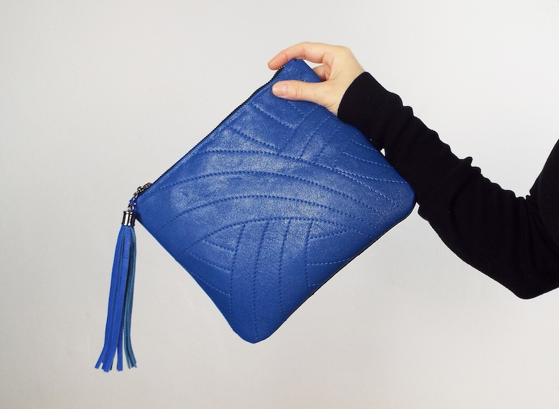 May include: A bright blue clutch bag with a quilted design. It has a matching blue tassel and a silver zip pull. The bag is set against a white backdrop.