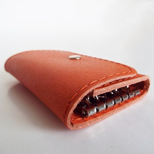 K&ouml;nnte beinhalten: Ein leuchtend orangefarbener Schl&uuml;sselanh&auml;nger aus Leder mit einem Druckknopfverschluss und einem Schl&uuml;sselring aus Metall. Der Schl&uuml;sselanh&auml;nger ist zusammengeklappt und der Schl&uuml;sselring ist sichtbar.