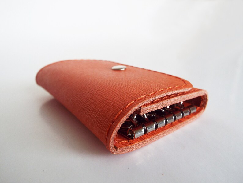 K&ouml;nnte beinhalten: Ein leuchtend orangefarbener Schl&uuml;sselanh&auml;nger aus Leder mit einem Druckknopfverschluss und einem Schl&uuml;sselring aus Metall. Der Schl&uuml;sselanh&auml;nger ist zusammengeklappt und der Schl&uuml;sselring ist sichtbar.