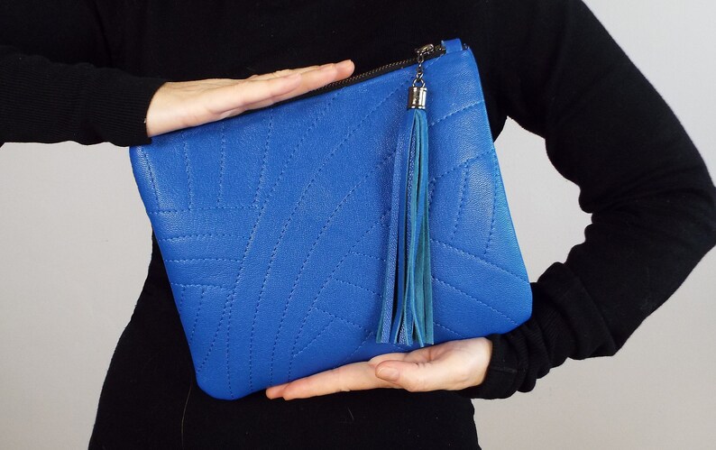 May include: A bright blue clutch bag with a stitched geometric design. It has a zip closure and a long, teal tassel. The bag is displayed against a plain background.