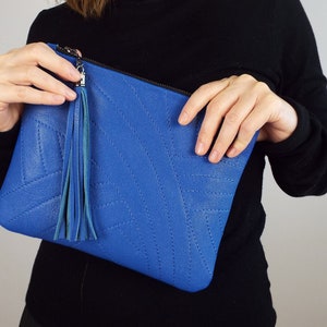 May include: A bright blue clutch bag with a stitched geometric design. It has a matching blue tassel and a silver zip. The bag is held by a person wearing a black long-sleeved top.