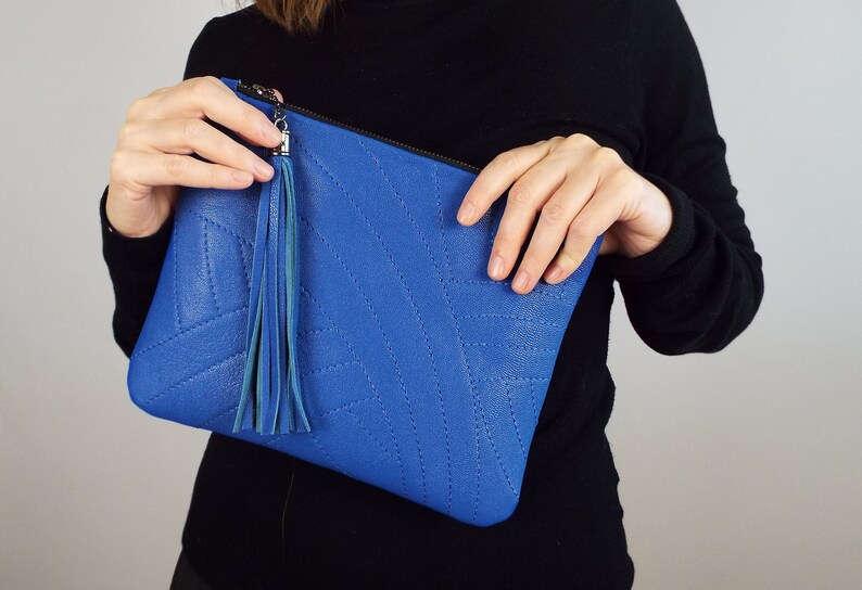 May include: A bright blue clutch bag with a stitched geometric design. It has a matching blue tassel and a silver zip. The bag is held by a person wearing a black long-sleeved top.