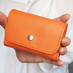 K&ouml;nnte beinhalten: Eine kleine orangefarbene Lederb&ouml;rse mit Druckknopfverschluss und einem silbernen Knopf.